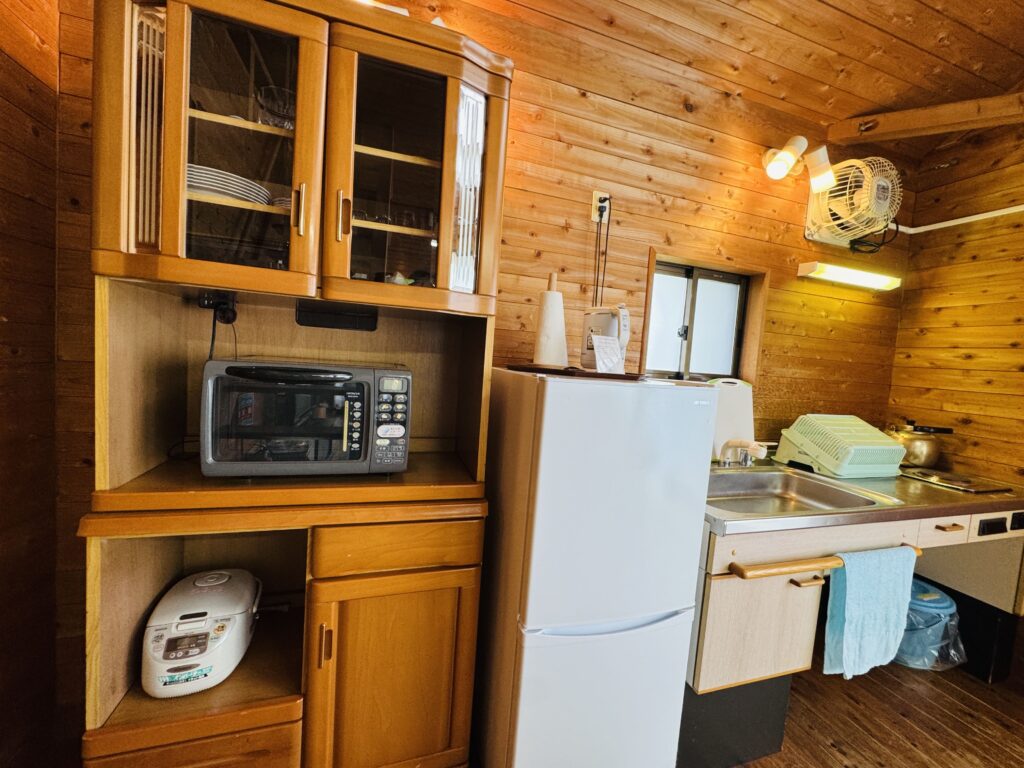 Kitchen area of a Kyukamura Fuji cottage featuring a refrigerator, microwave, rice cooker, and other basic cooking appliances.