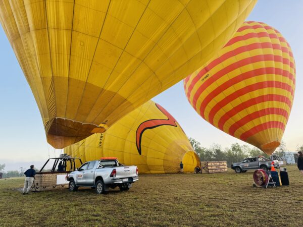 Hot air balloons being prepared for takeoff in Mareeba, near Cairns, just before sunrise. The balloons slowly inflate under the quiet pre-dawn sky.
