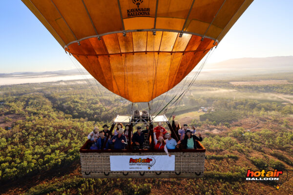 Group photo taken by an external camera mounted on the hot air balloon, showing the entire balloon, smiling passengers, and the glowing sunrise sky over Cairns.